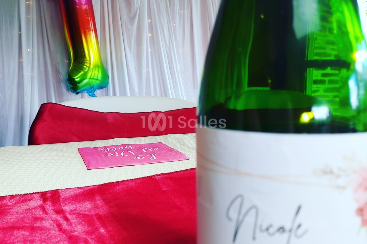 Ballon arc-en-ciel en forme de chiffre un, table décorée avec nappe rouge et bouteille au premier plan.