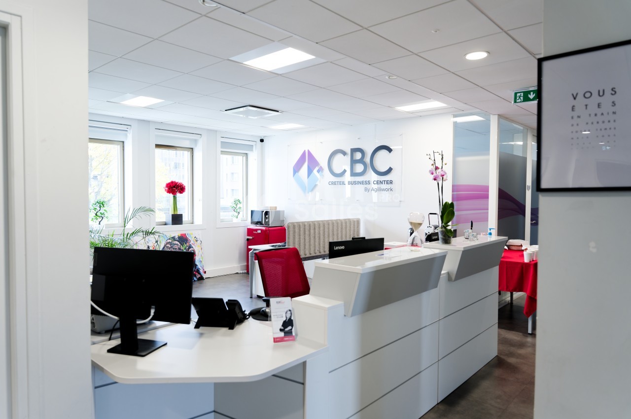 Espace d'accueil moderne avec comptoir blanc, chaises rouges, plantes et logo ’CBC’ sur un mur vitré.