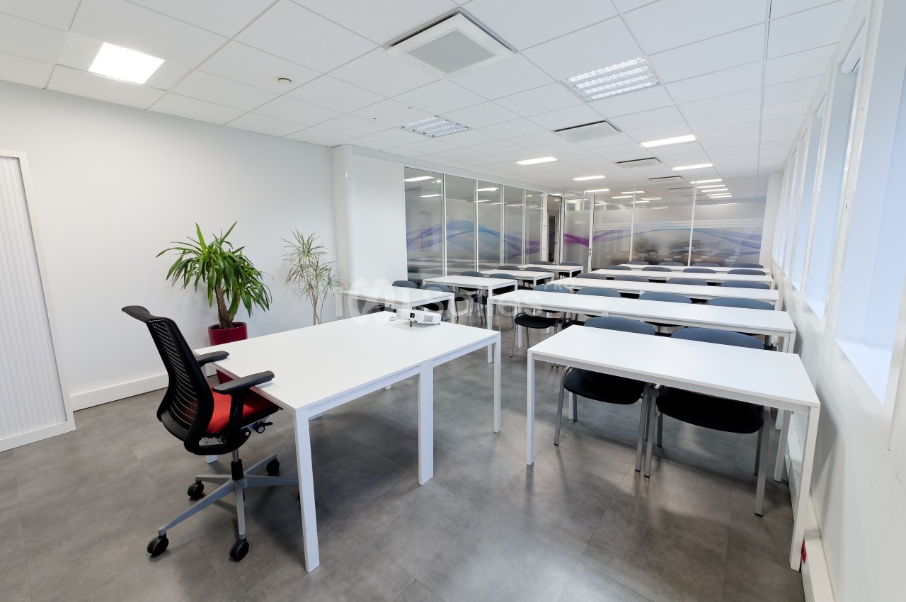 Salle de réunion lumineuse avec tables blanches, chaises noires, plantes et grandes baies vitrées.