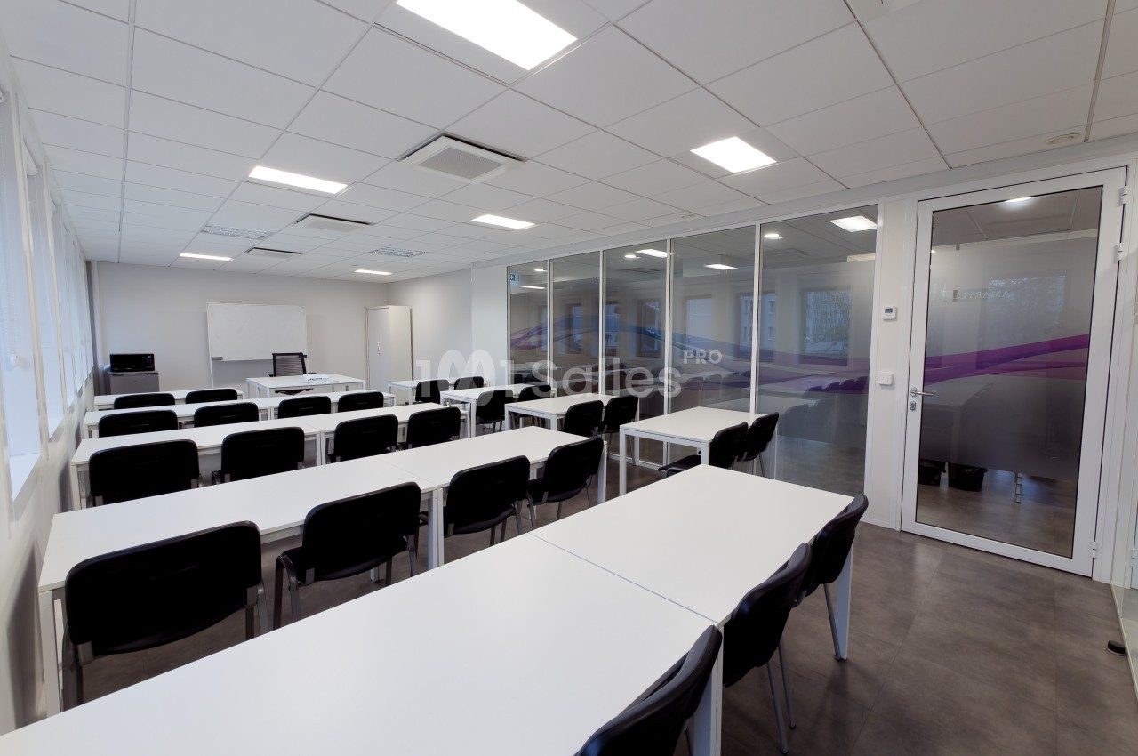 Salle de formation lumineuse avec des tables blanches, des chaises noires et un tableau blanc au fond.