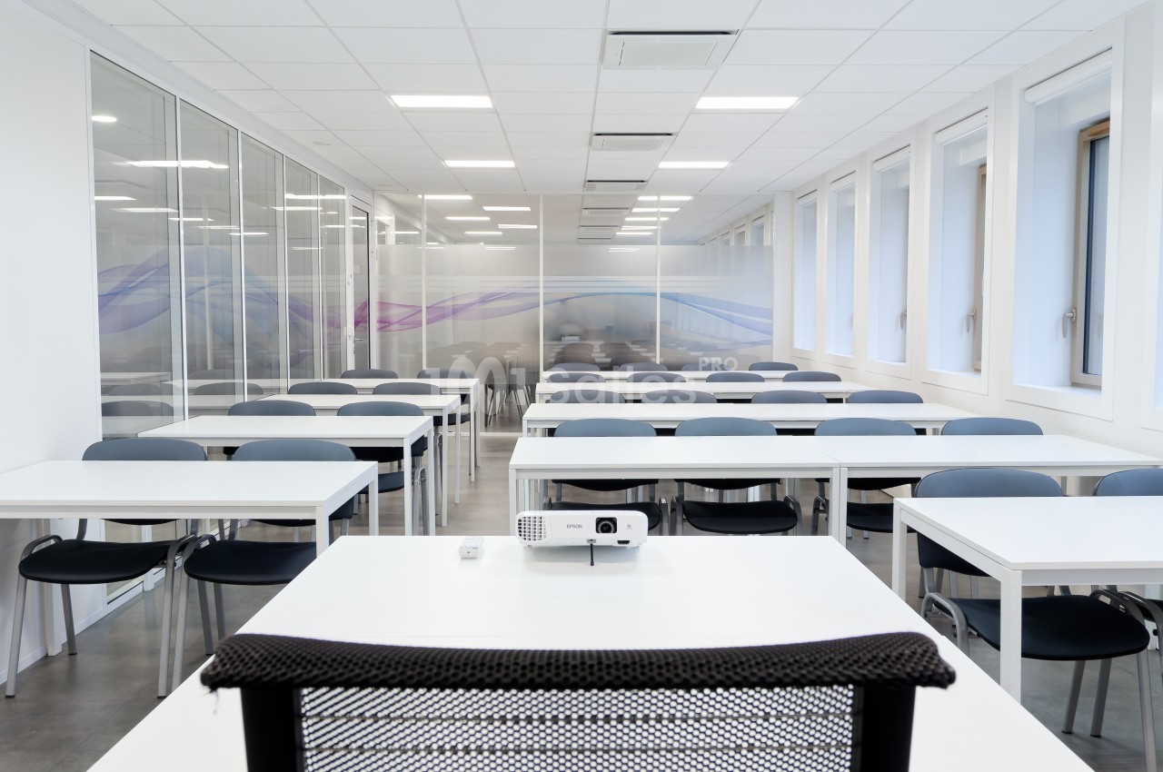Salle de classe moderne et lumineuse avec des rangées de tables et chaises, vue depuis un bureau équipé d’un projecteur.