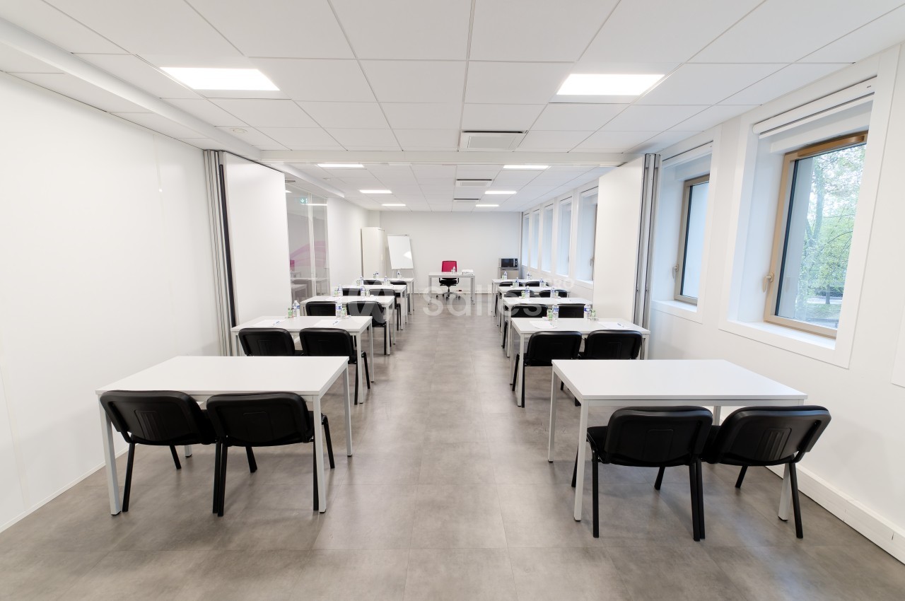 Salle de formation lumineuse avec tables blanches, chaises noires et grandes fenêtres laissant entrer la lumière naturelle.