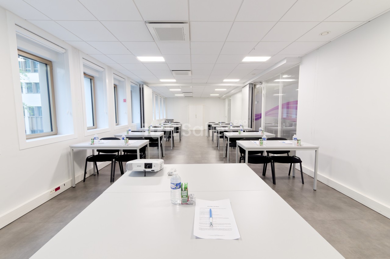 Salle de formation lumineuse avec des tables alignées, des chaises, des documents et une bouteille d'eau sur chaque table.