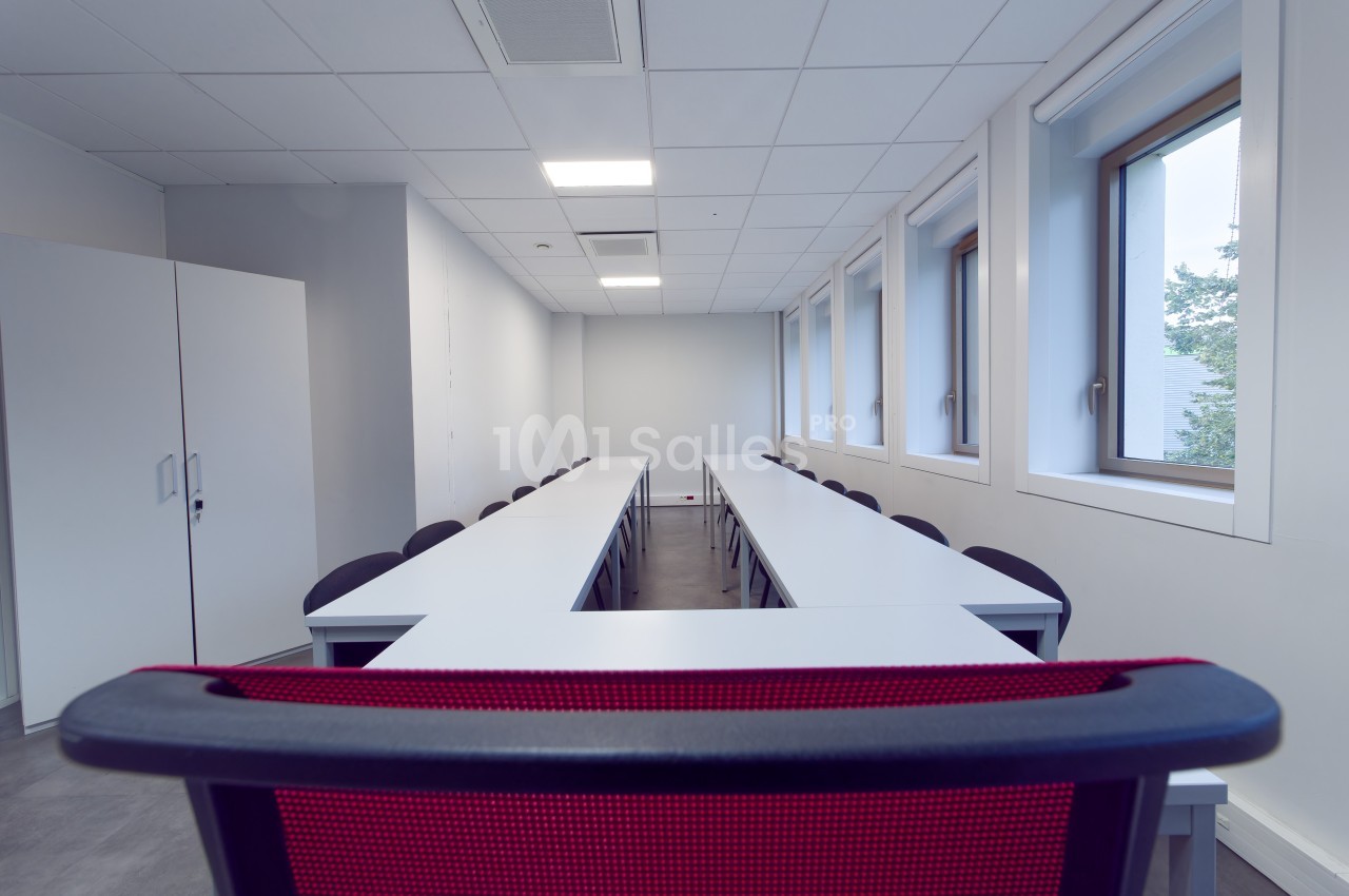 Salle de réunion vide avec tables blanches disposées en U, chaises noires et fenêtres laissant entrer la lumière naturelle.
