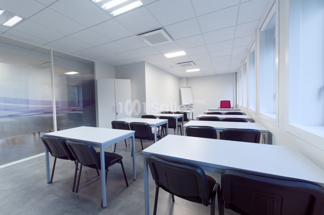 Salle de classe vide avec des tables, des chaises noires, un tableau blanc et de grandes fenêtres laissant entrer la lumière.