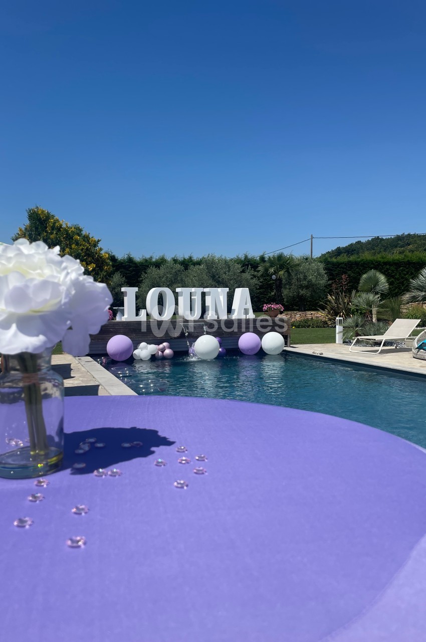 Table violette décorée de perles et fleurs blanches, près d'une piscine entourée de ballons et d'une végétation estivale.