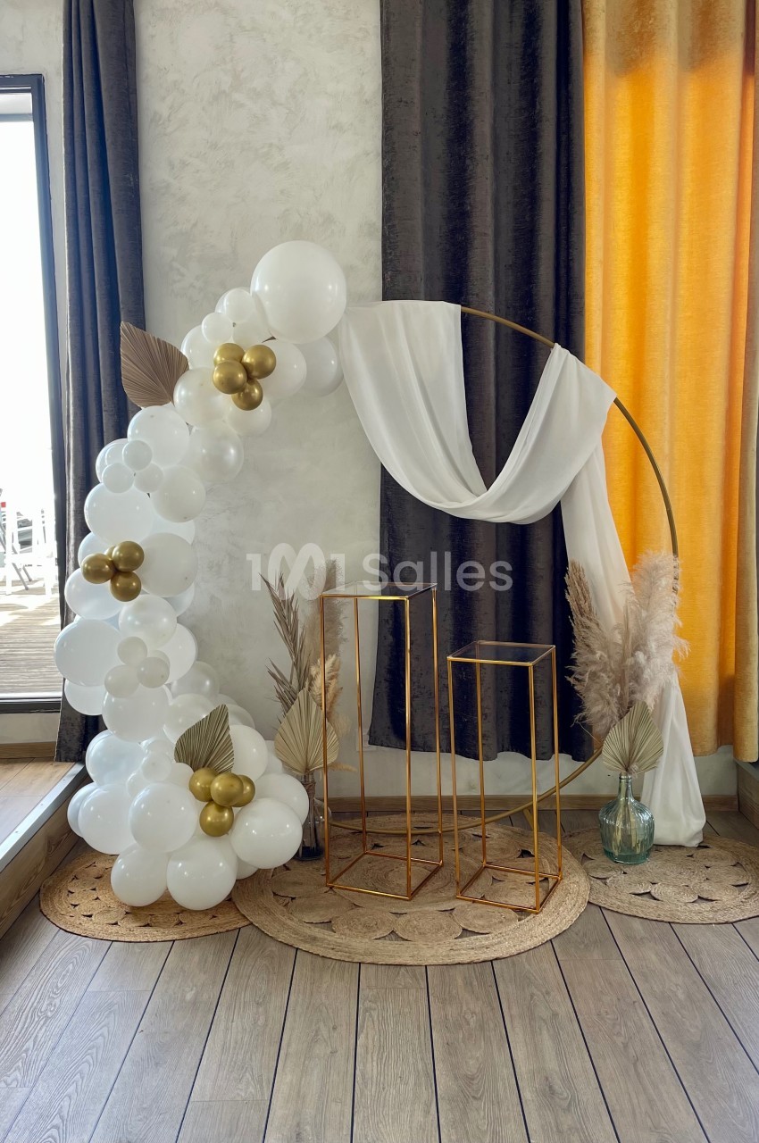 Décor élégant avec arche dorée, rideaux, ballons blancs et dorés, et éléments végétaux dans un cadre intérieur lumineux.