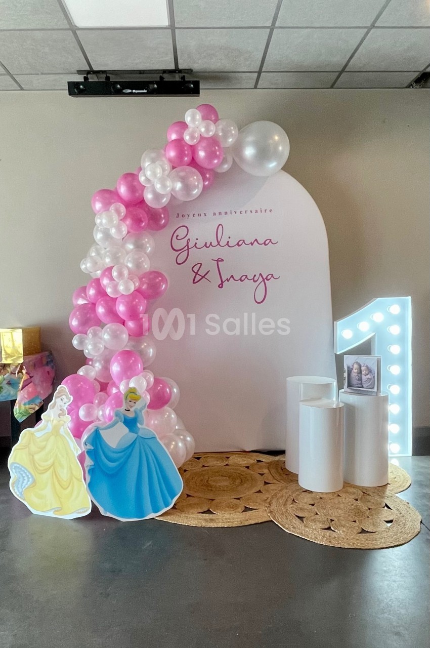 Décor d'anniversaire avec arche de ballons roses et blancs, silhouettes de princesses et chiffre lumineux ’1’.