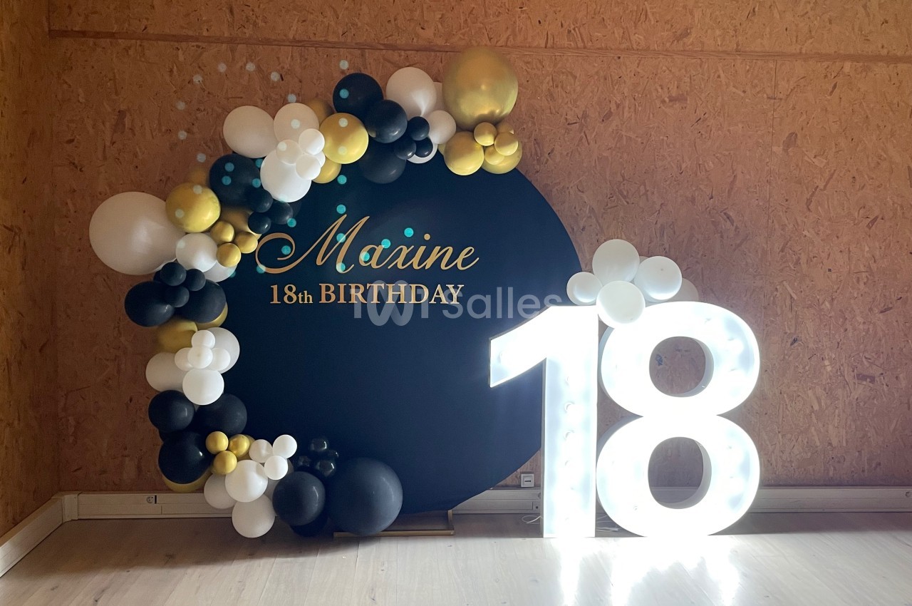 Décoration d'anniversaire avec un panneau noir orné de ballons et un chiffre lumineux ’18’ en blanc.