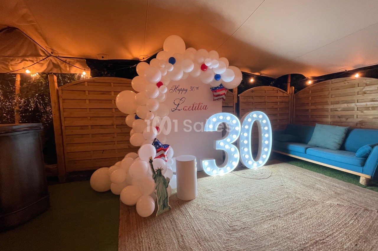 Décoration d'anniversaire avec ballons blancs, chiffre lumineux ’30’ et guirlandes lumineuses dans un espace couvert.