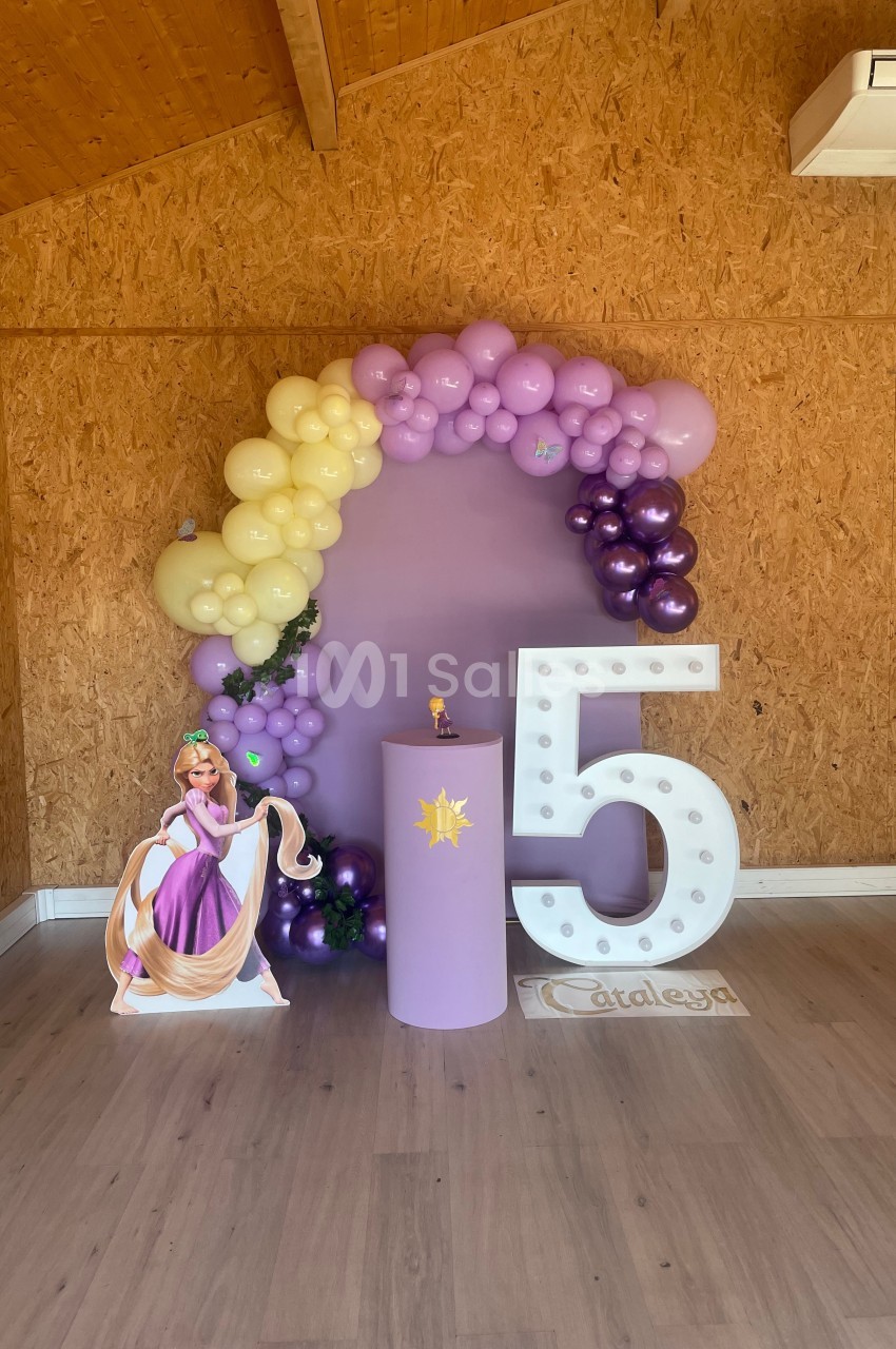 Décoration d'anniversaire avec ballons violets et jaunes, chiffre 5 lumineux, et figurine de princesse devant un mur en bois.