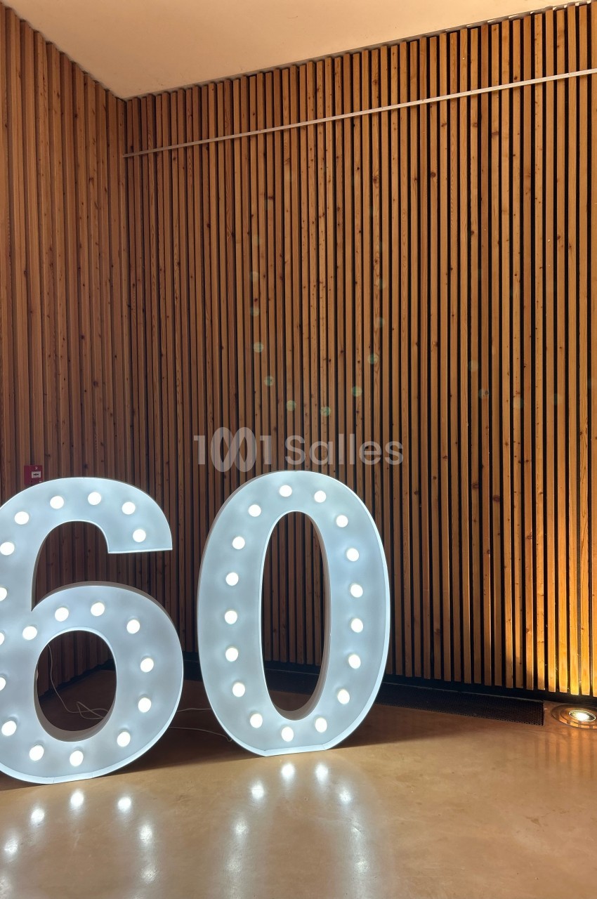Chiffres lumineux ’60’ posés sur un sol en bois, devant un mur en lambris vertical.