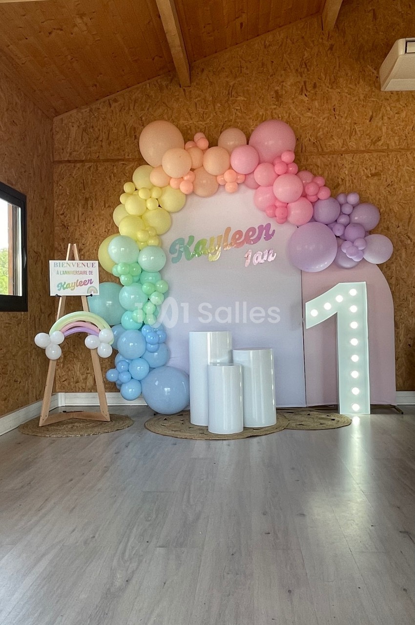 Décoration d'anniversaire avec arche de ballons pastel, chiffre lumineux ’1’ et panneau personnalisé dans une salle en bois.
