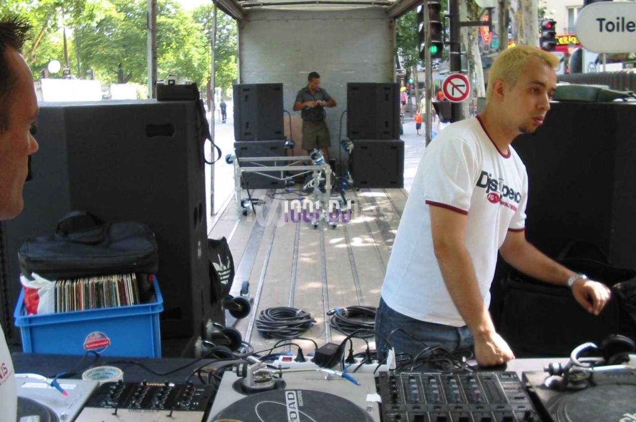 Un DJ prépare son équipement sur une scène extérieure avec des platines et des enceintes en arrière-plan.