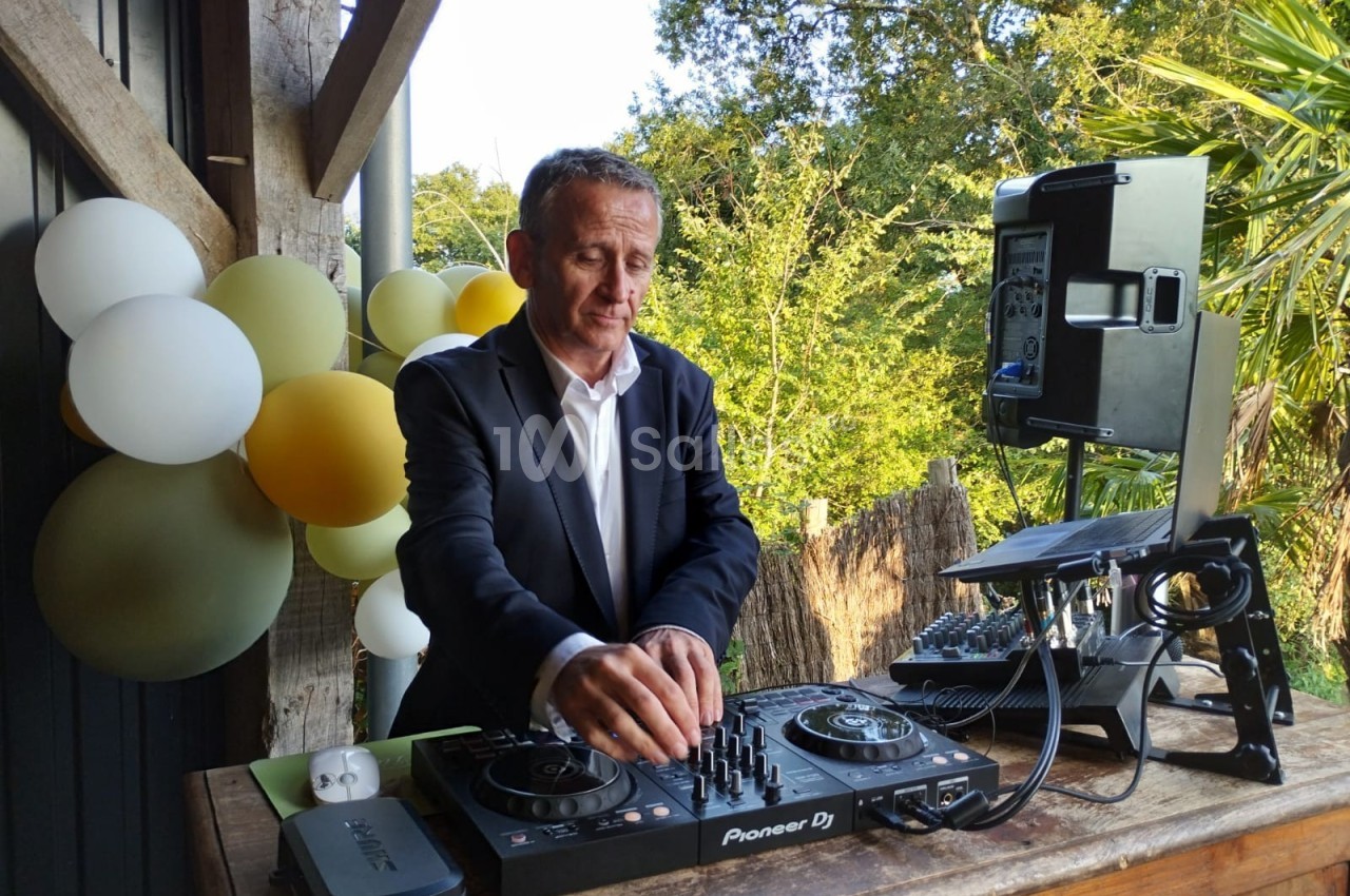Un homme en costume mixe de la musique sur une table de DJ en extérieur, avec des ballons et de la verdure en arrière-plan.