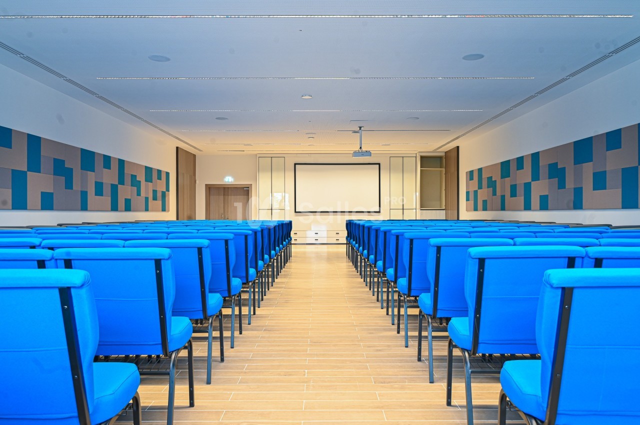 Salle de conférence vide avec des rangées de chaises bleues, un écran de projection et des panneaux acoustiques au mur.