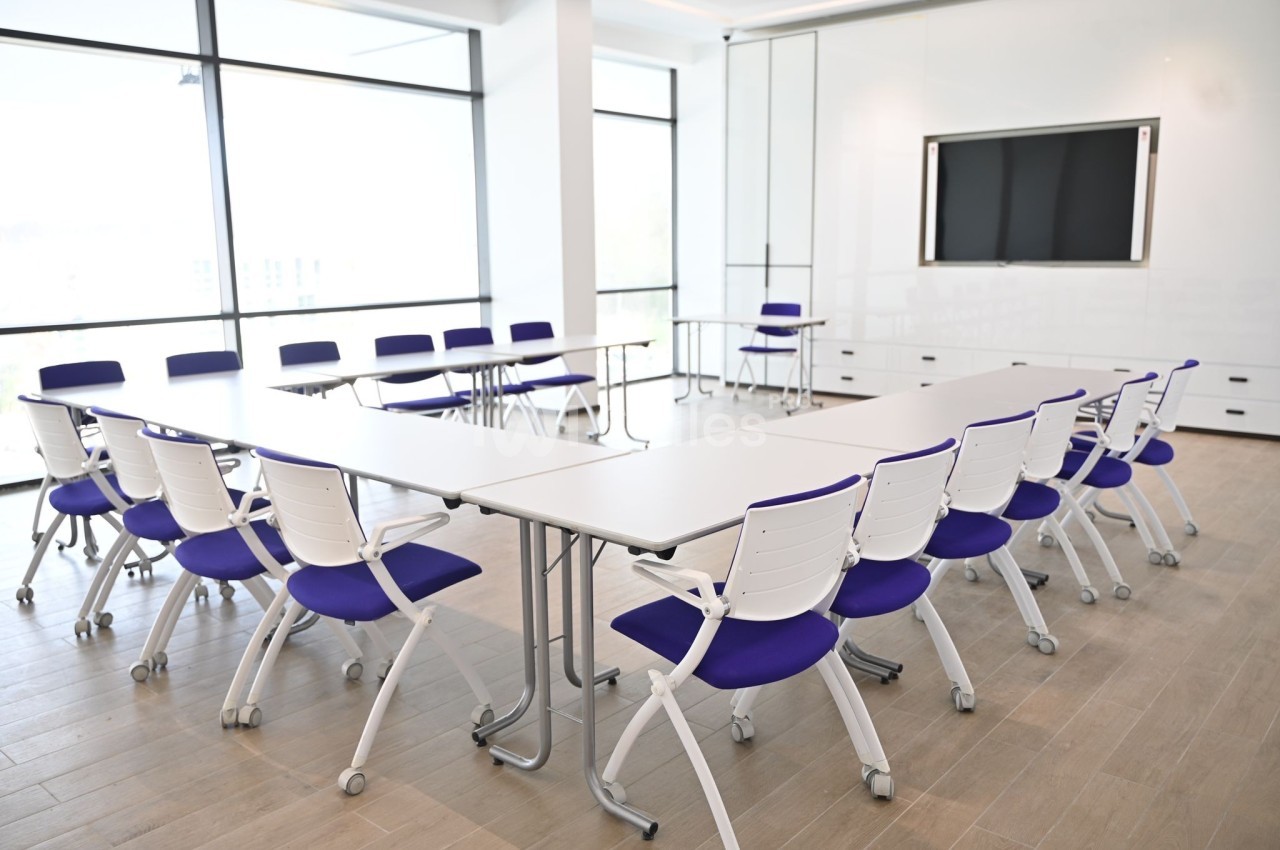 Salle de réunion lumineuse avec tables blanches disposées en U et chaises à assise violette sur un sol en bois clair.