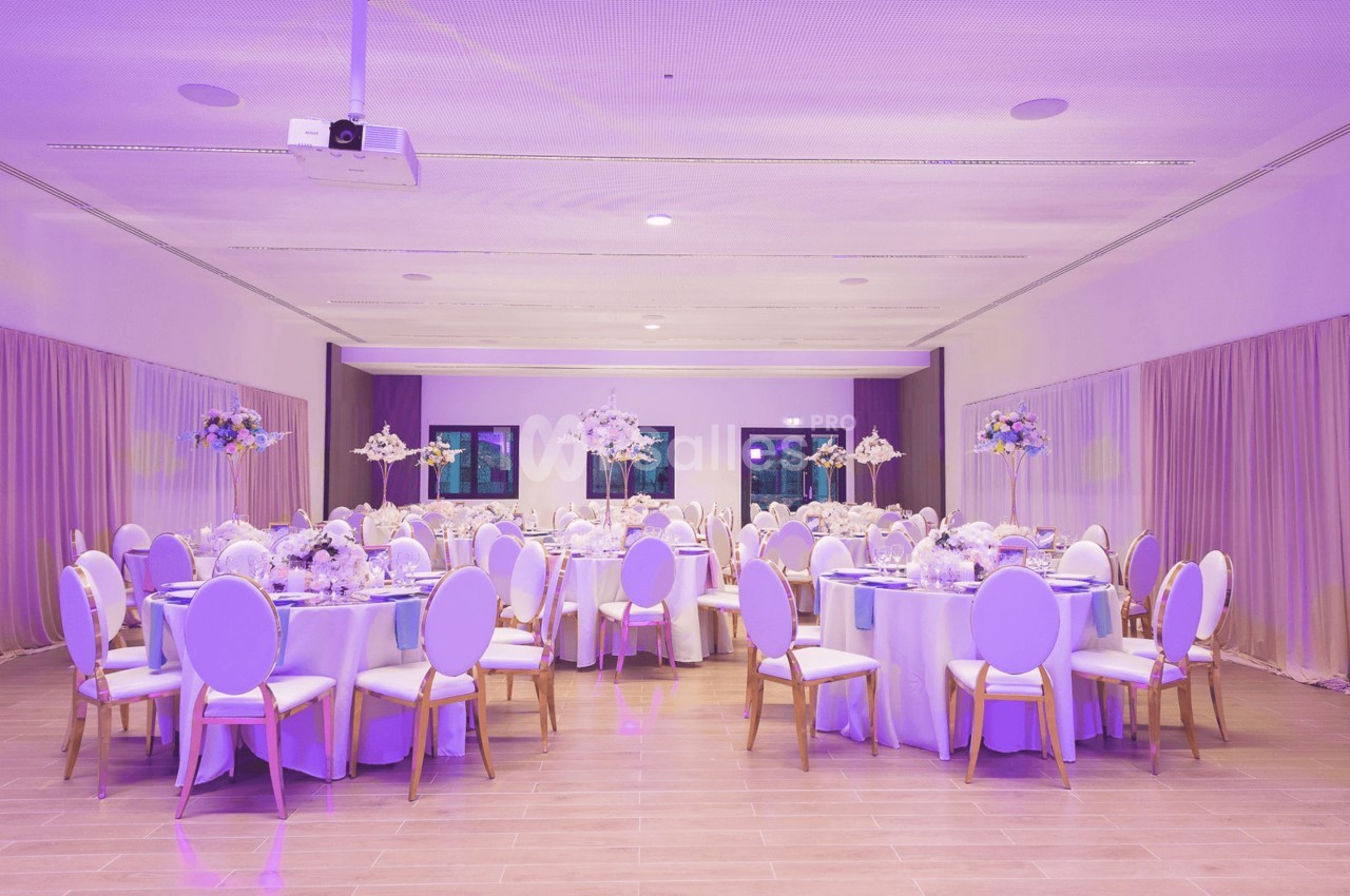 Salle de réception décorée avec des tables rondes, nappes blanches, chaises élégantes et éclairage violet.