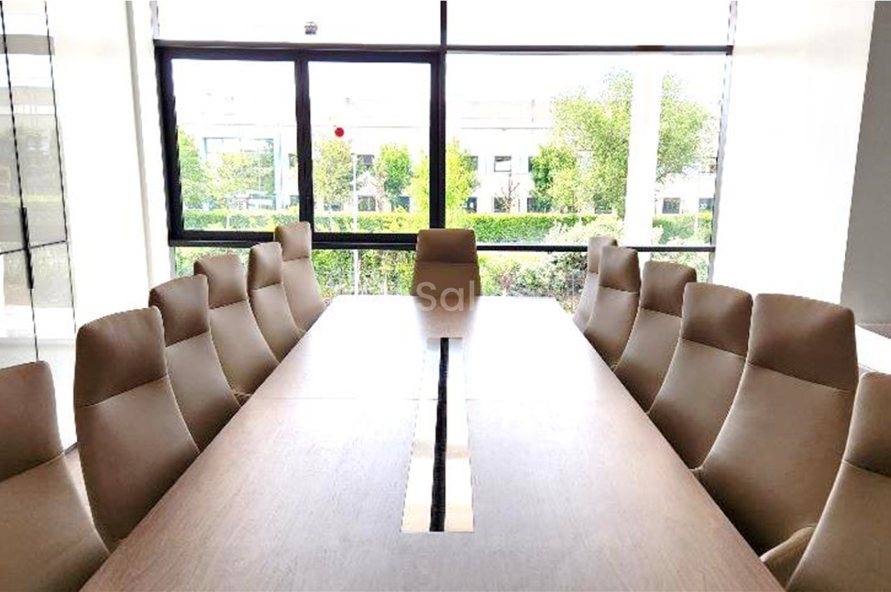 Salle de réunion lumineuse avec une grande table en bois entourée de chaises beige, vue sur des fenêtres donnant sur un…