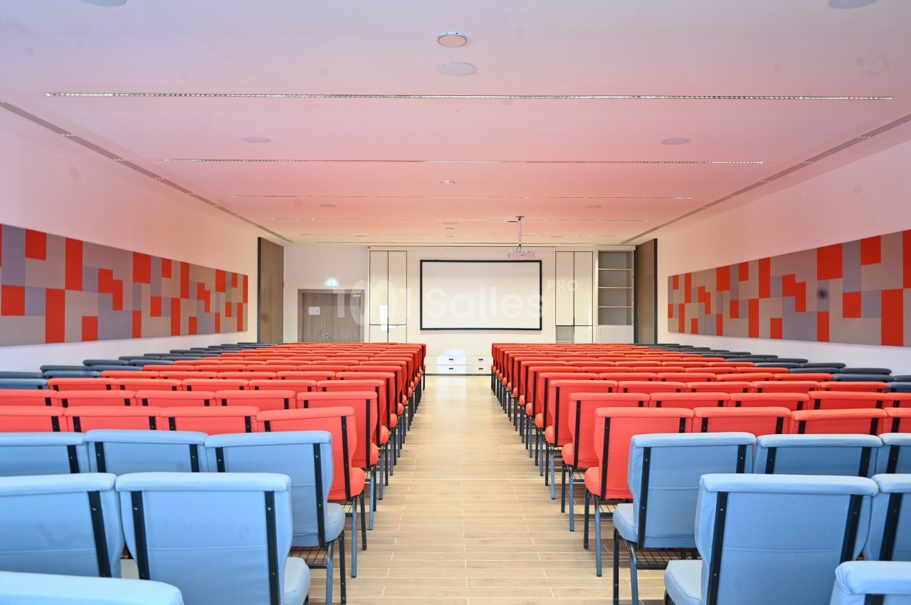 Salle de conférence moderne avec rangées de sièges rouges et bleus, écran de projection et murs décorés de panneaux colorés.