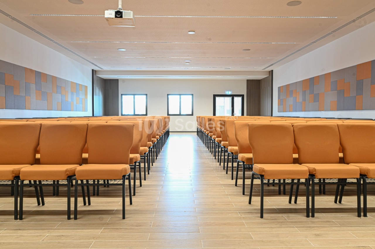 Salle de conférence lumineuse avec des rangées de chaises orange alignées face à un mur équipé d'un projecteur.