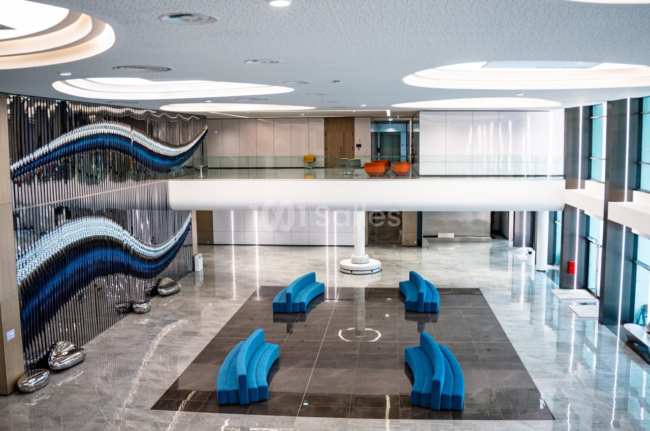 Hall moderne avec sol en marbre, sièges bleus courbés, sculptures murales ondulées et mezzanine vitrée.
