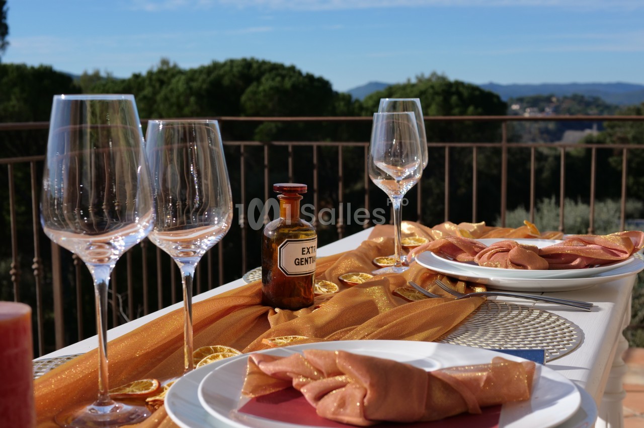 Table extérieure élégamment dressée avec nappes dorées, verres à vin et vue sur un paysage arboré et montagneux.