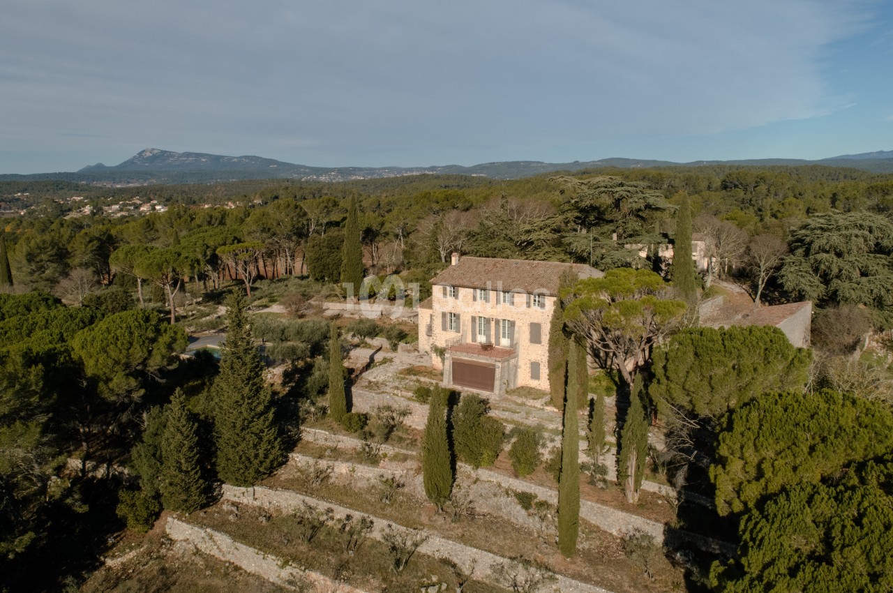 Maison en pierre entourée de terrasses arborées et de cyprès, située dans un paysage naturel avec collines en arrière-plan.