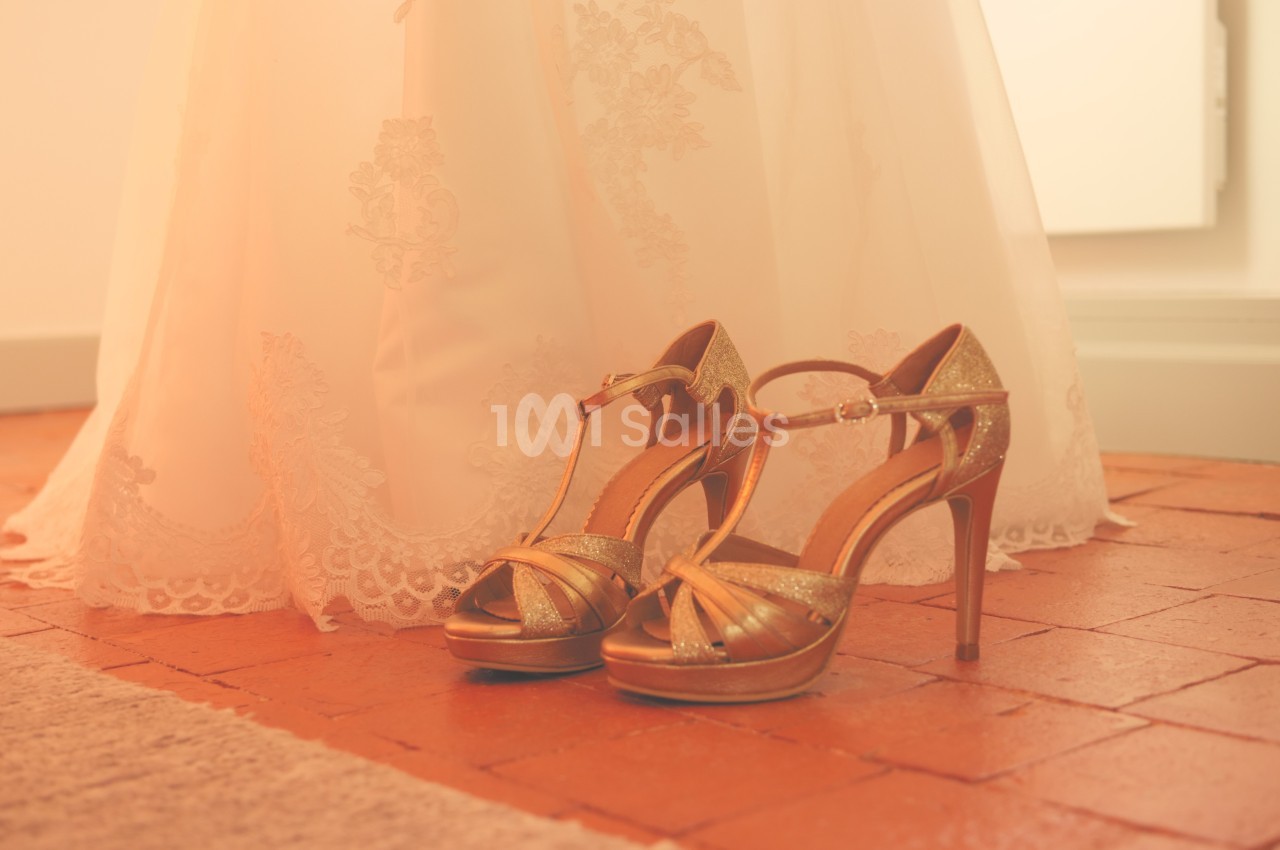 Chaussures à talons dorées posées sur un sol en terre cuite, devant une robe en dentelle blanche.