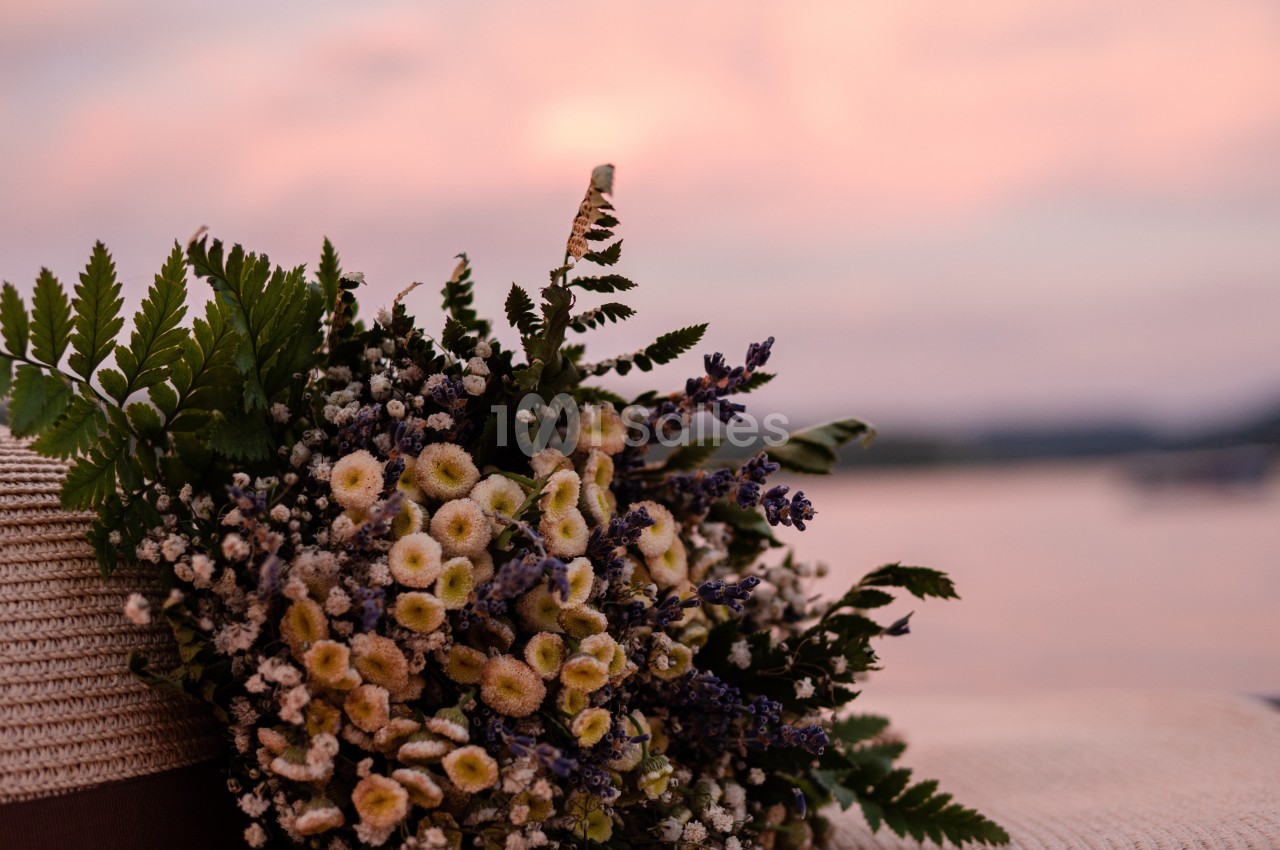 Bouquet de fleurs sauvages posé sur un chapeau, avec un ciel rose en arrière-plan au bord de l'eau.