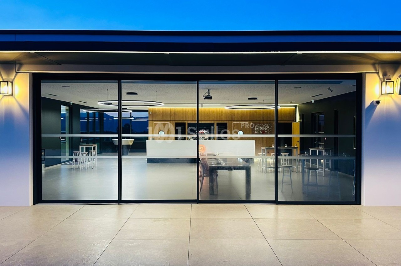 Façade vitrée d'un bâtiment moderne éclairé, donnant sur un espace intérieur avec mobilier minimaliste.
