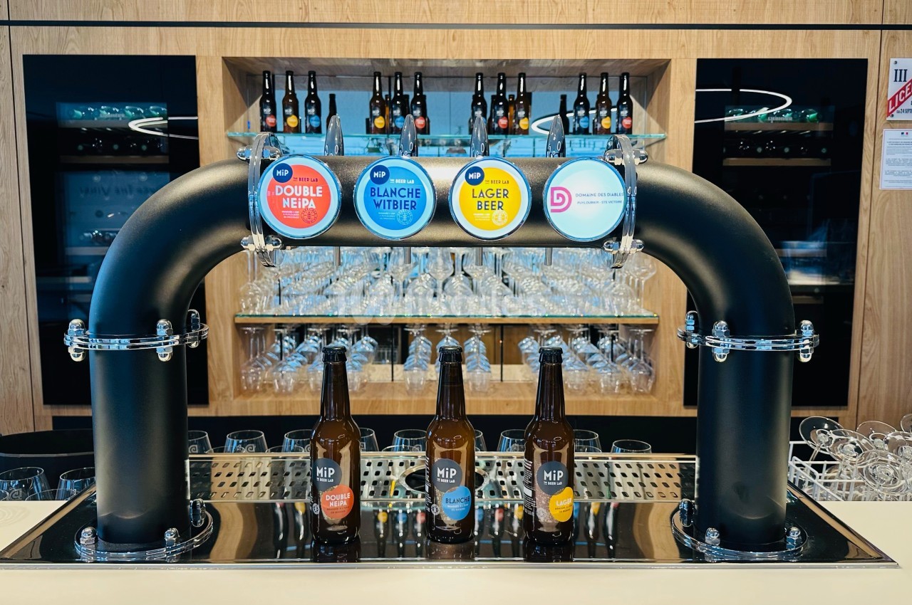 Tireuse à bière avec quatre robinets et bouteilles alignées devant, dans un bar moderne avec étagères de verres.