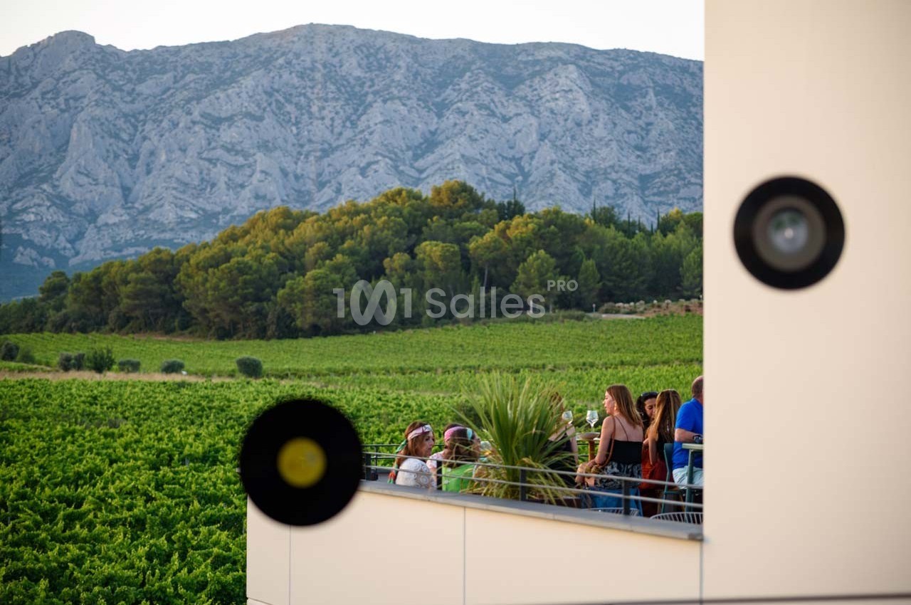 Des personnes discutent sur une terrasse surplombant un vignoble verdoyant avec des montagnes en arrière-plan.