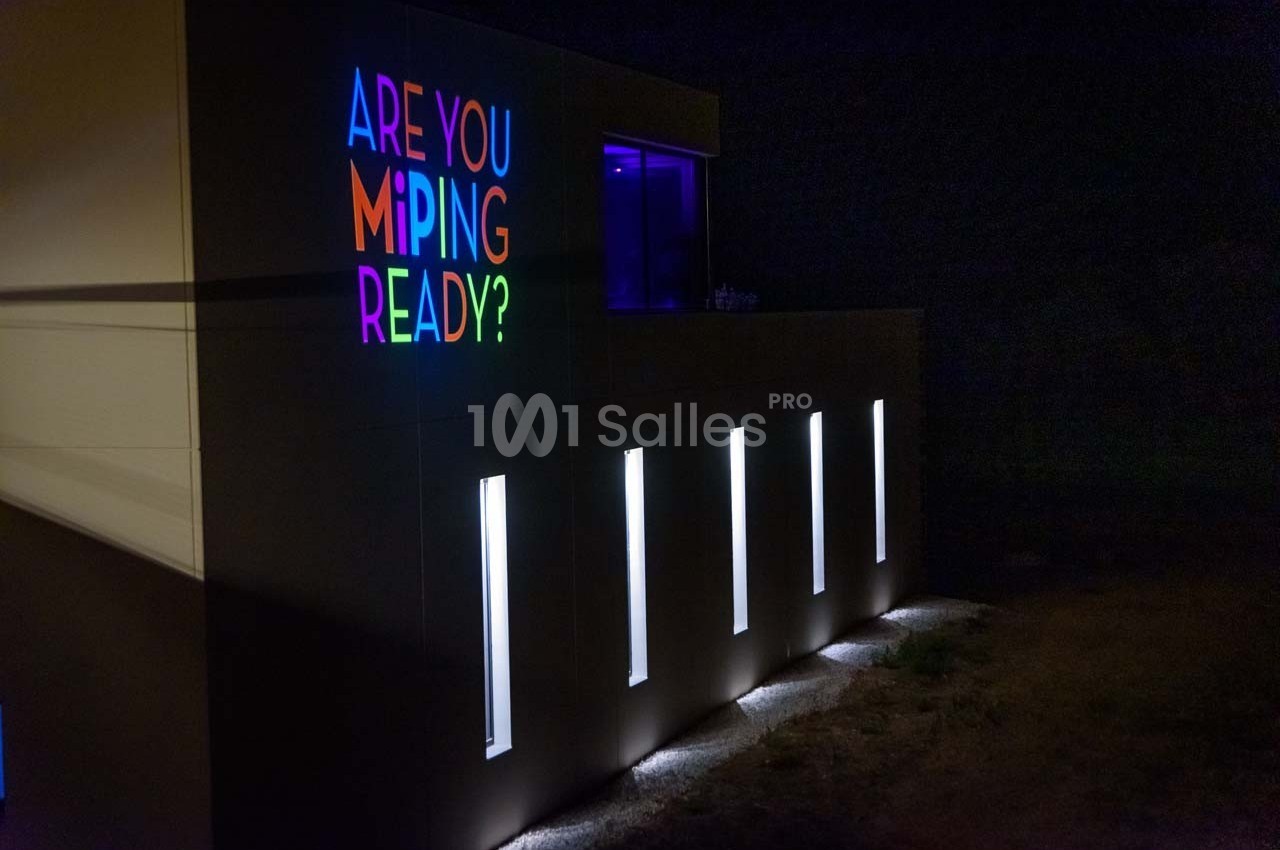 Façade d'un bâtiment moderne éclairée la nuit, avec un message coloré ’Are you Miping ready?’ sur le mur.