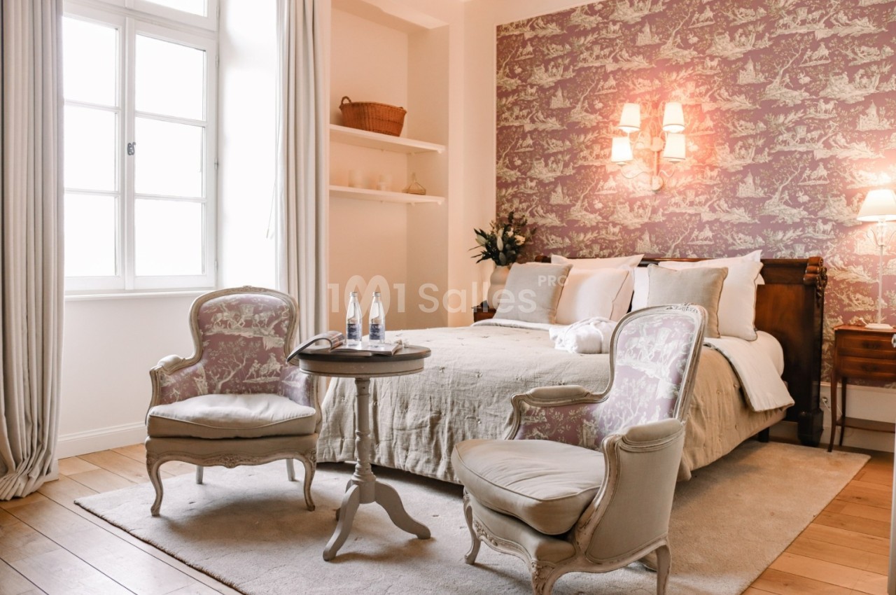 Chambre lumineuse avec lit double, fauteuils anciens, table basse, mur décoré de papier peint et fenêtre à gauche.