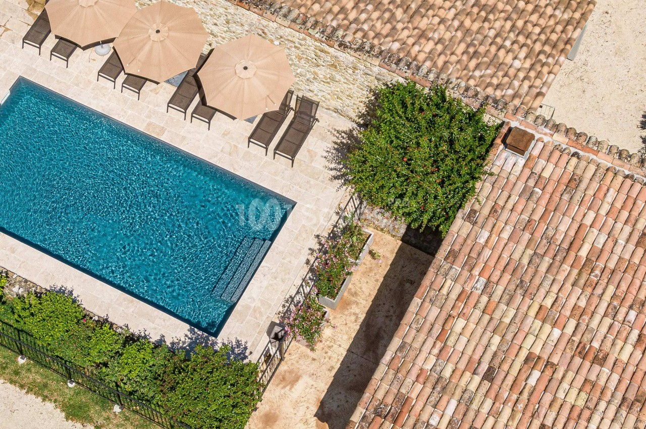Vue aérienne d'une piscine entourée de chaises longues, parasols, végétation et bâtiments en tuiles.