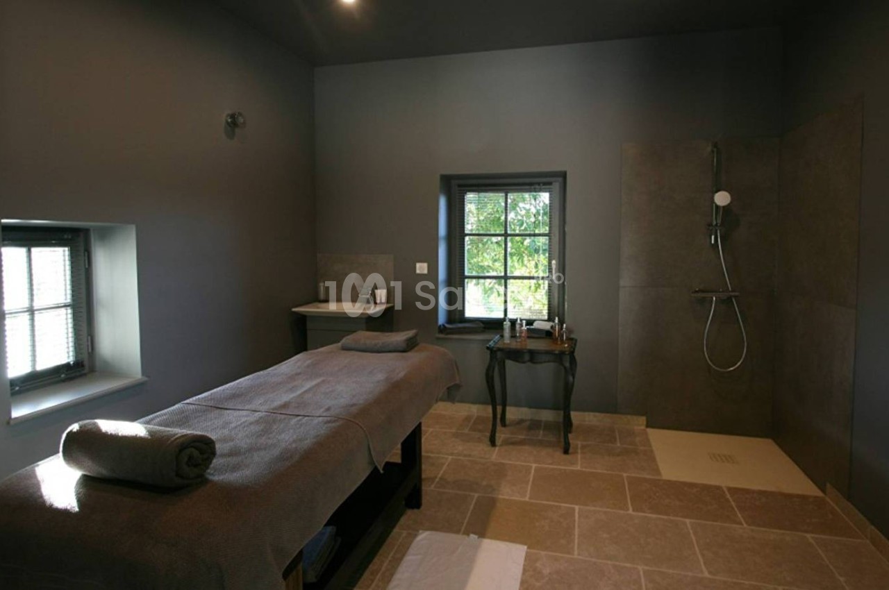 Salle de massage avec table recouverte d'une serviette, douche à l'italienne et lumière naturelle entrant par les fenêtres.