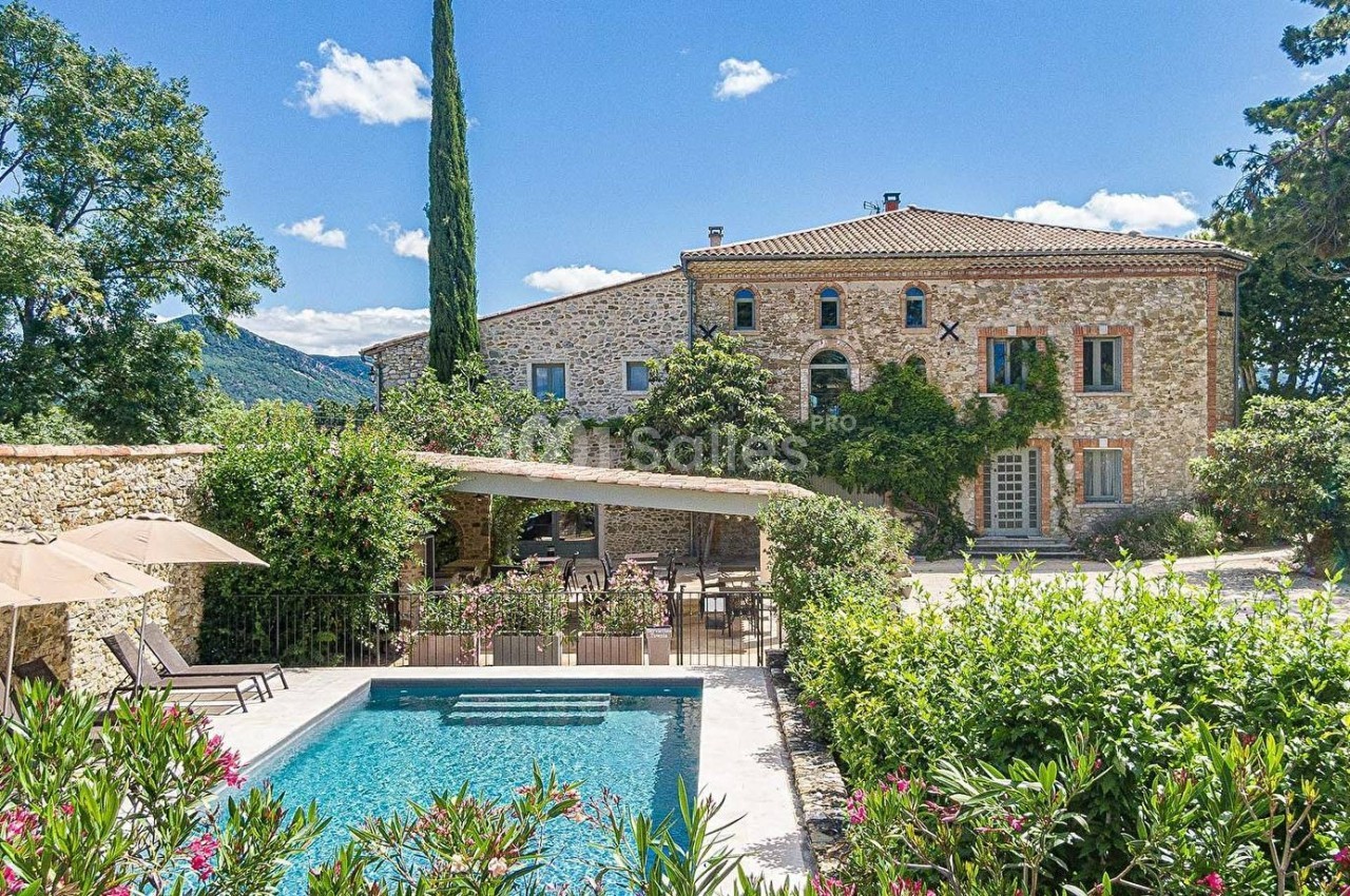 Maison en pierre entourée de verdure avec une piscine et une terrasse ombragée, sous un ciel bleu clair.