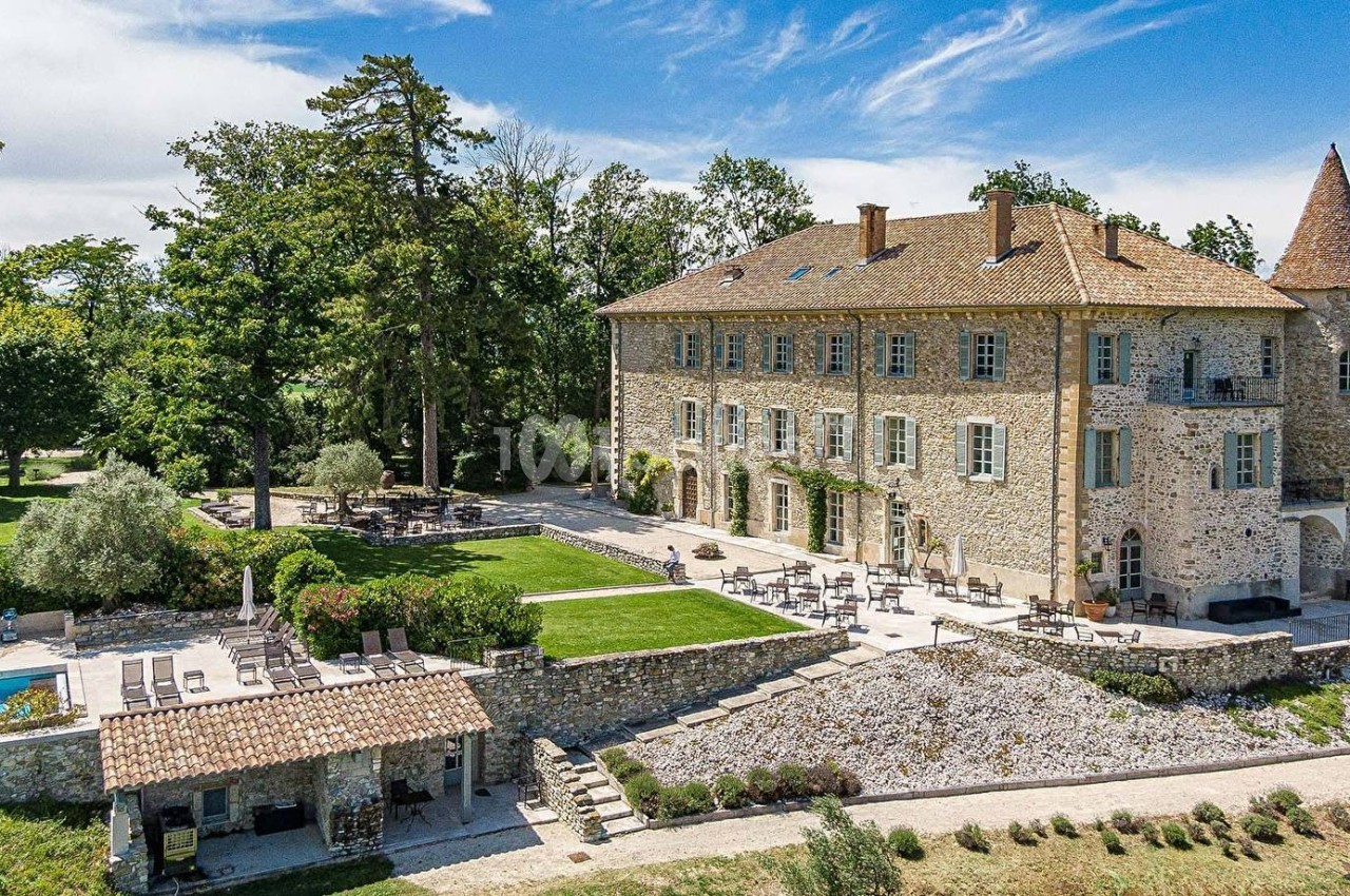 Vue d'un grand bâtiment en pierre entouré de jardins aménagés, avec une terrasse et un ciel dégagé.