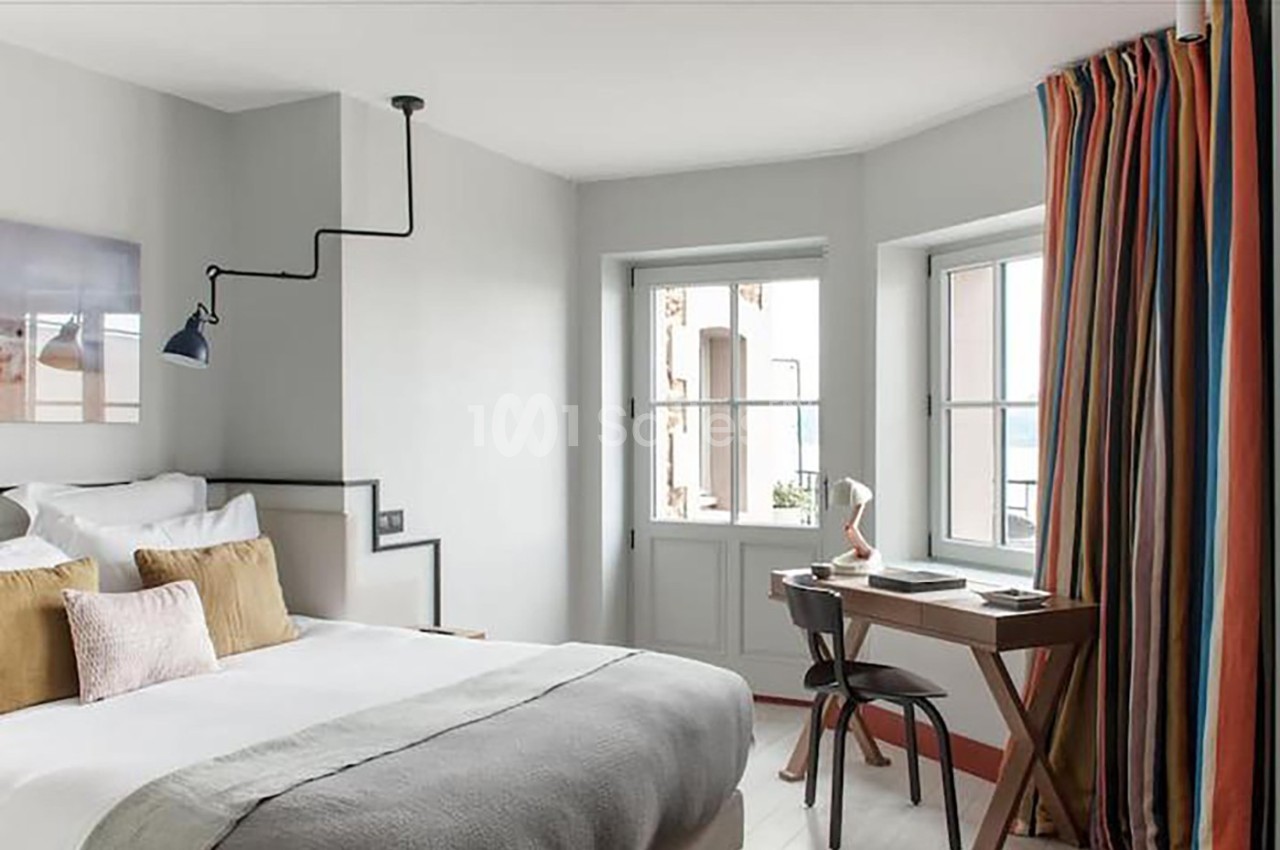 Chambre lumineuse avec lit double, bureau en bois, lampe et rideaux colorés près de fenêtres donnant sur l'extérieur.