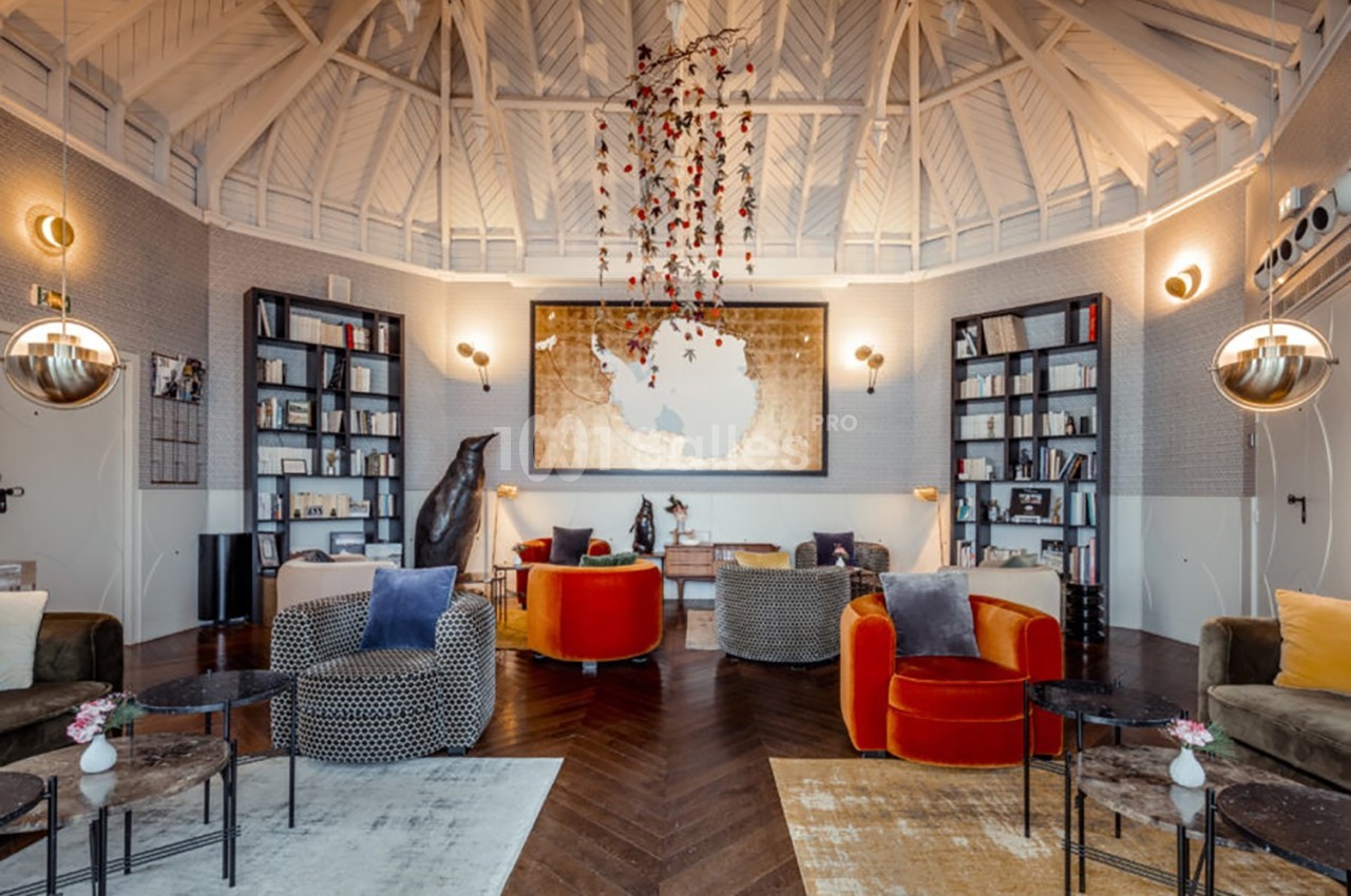 Salon lumineux avec fauteuils colorés, tables basses, étagères remplies de livres et plafond voûté en bois blanc.