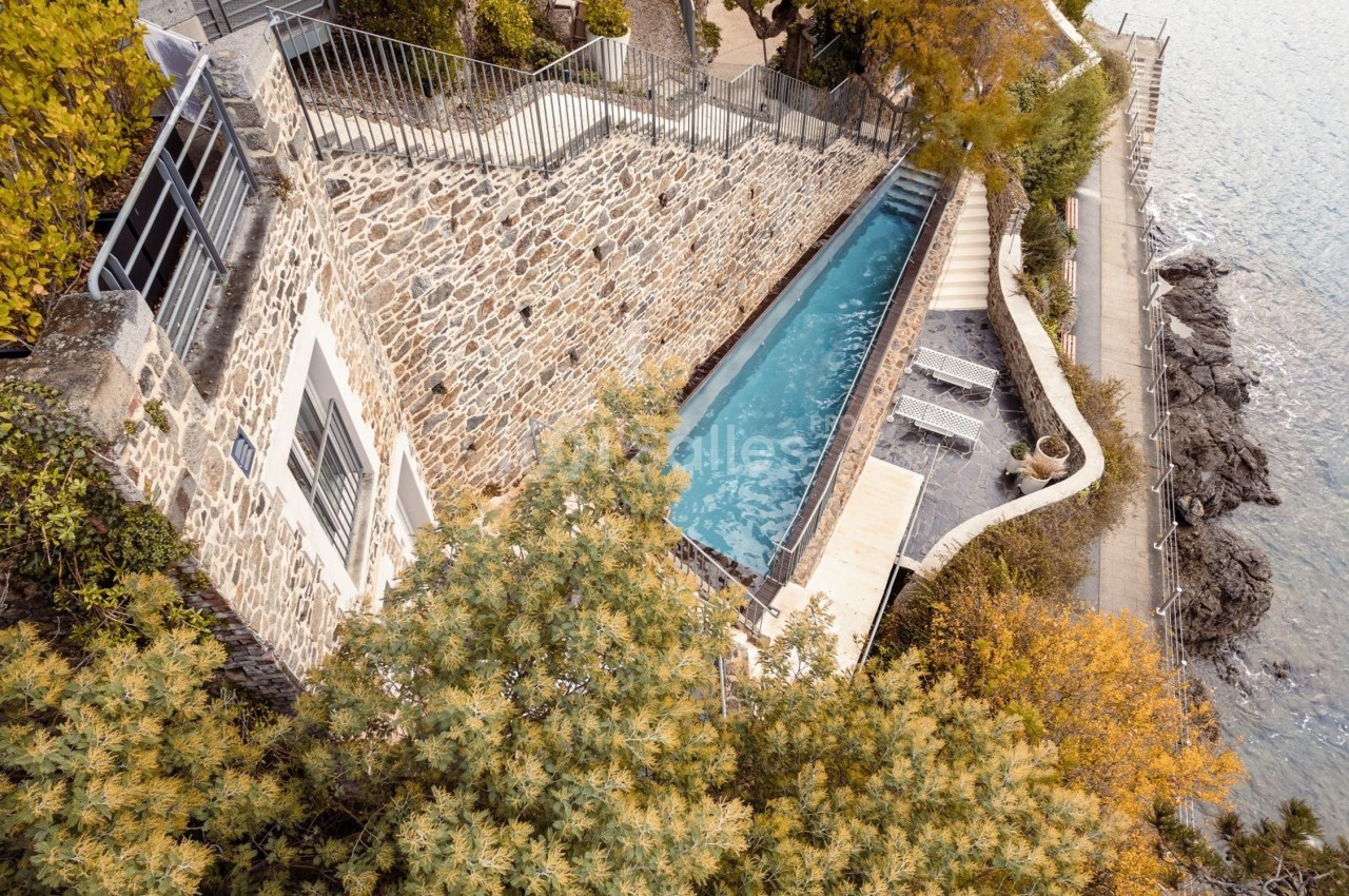 Piscine étroite entourée de murs en pierre, située en bord de mer avec végétation et chemin adjacent.