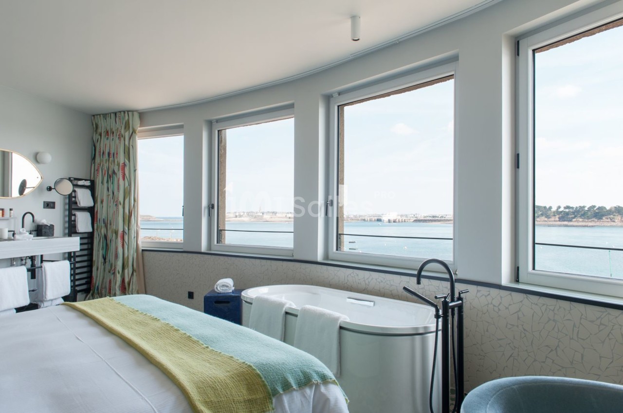 Chambre lumineuse avec lit, baignoire et vue sur la mer à travers de grandes fenêtres.