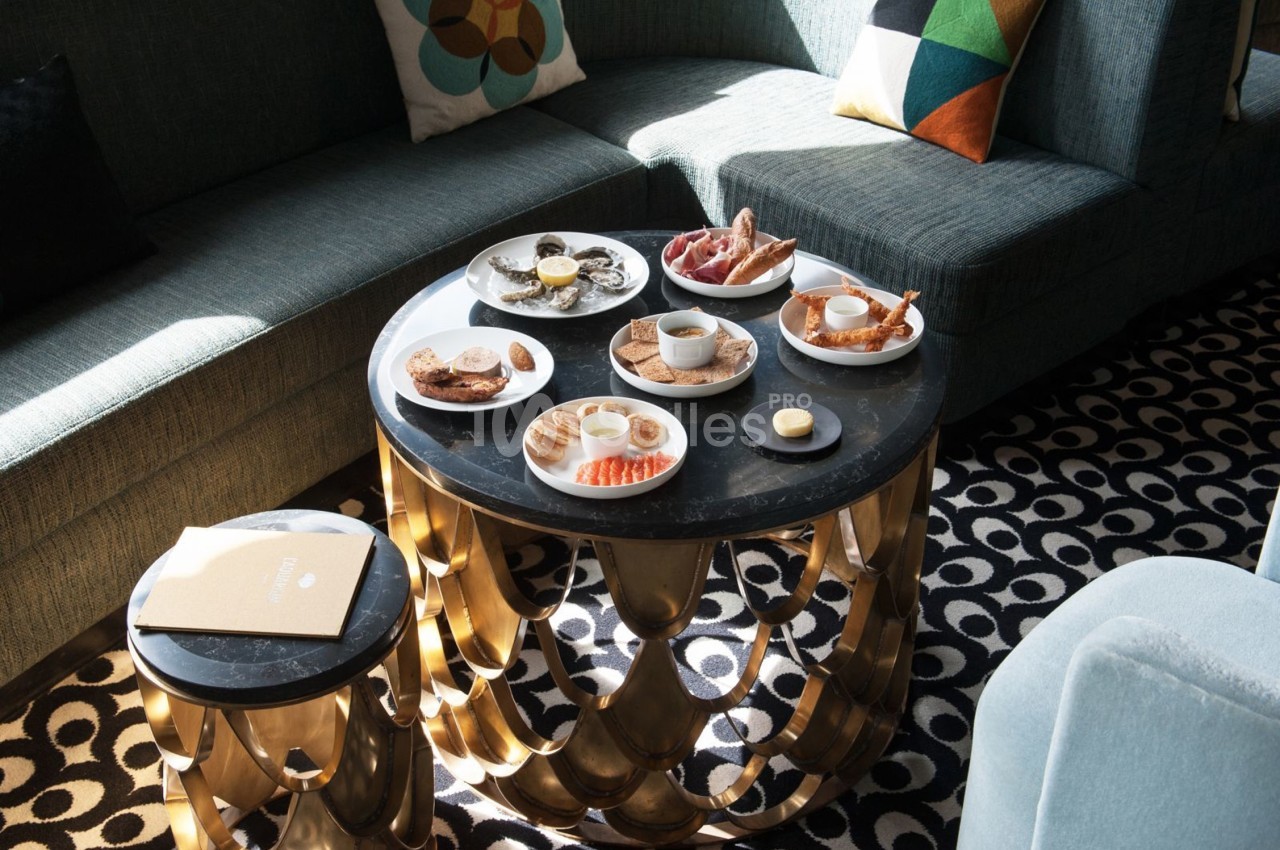 Table basse ronde avec petit-déjeuner varié, comprenant viennoiseries, fruits et boissons, dans un salon lumineux.