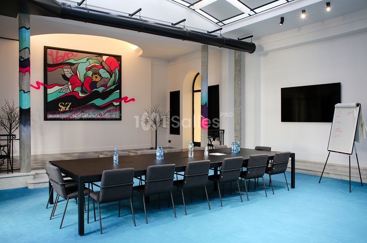 Salle de réunion moderne avec table noire, chaises, tableau blanc, écran mural et décoration artistique colorée.