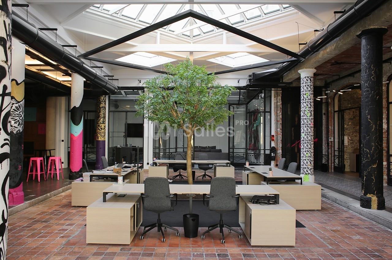 Espace de travail moderne avec bureaux partagés, un arbre central et une verrière laissant entrer la lumière naturelle.