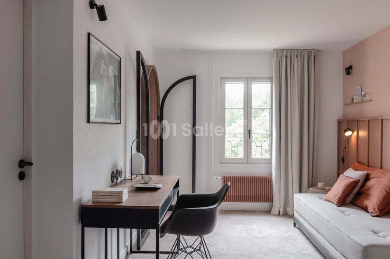 Bureau avec chaise noire, lampe et livres, dans une chambre lumineuse avec canapé, rideaux clairs et fenêtre ouverte.
