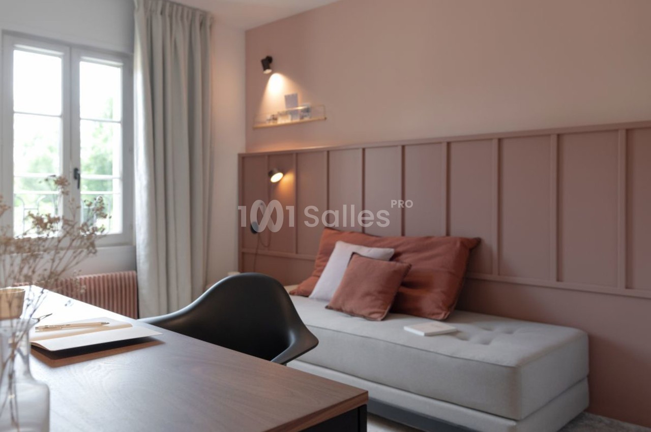Chambre lumineuse avec un lit gris, coussins terracotta, bureau en bois et mur rose décoré de moulures.