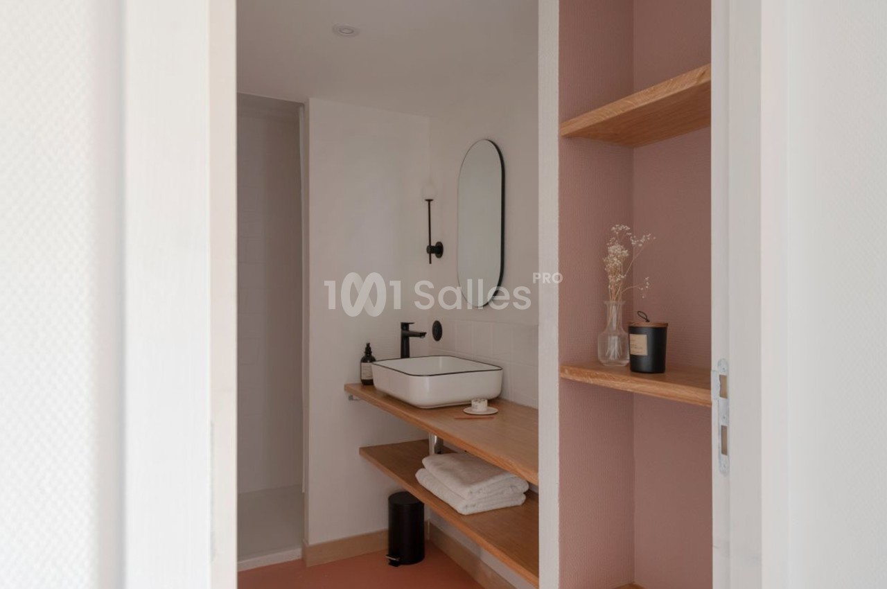 Salle de bain minimaliste avec lavabo blanc, miroir ovale, étagères en bois et mur rose pâle.