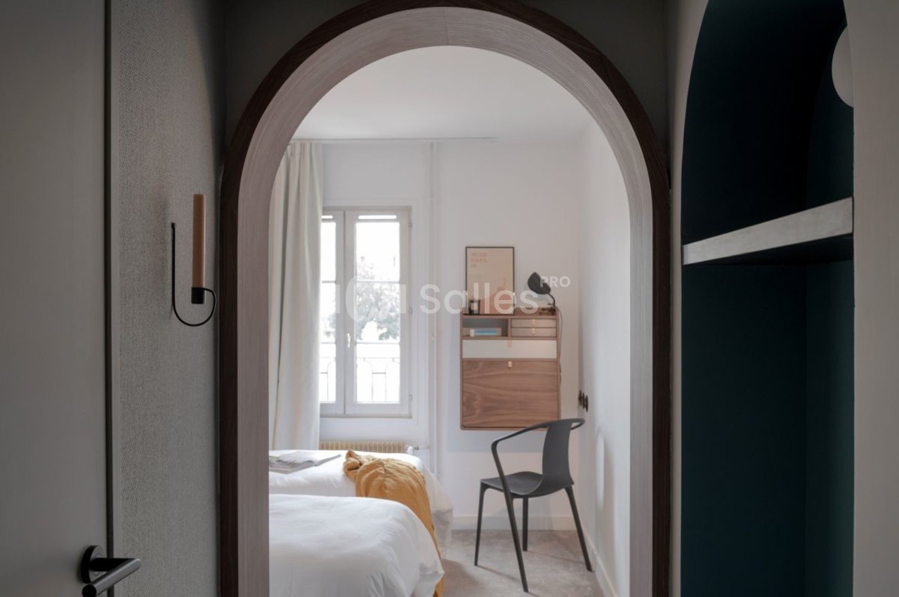 Vue d'une chambre lumineuse avec un lit, une chaise, un bureau mural en bois et une fenêtre encadrée de rideaux clairs.